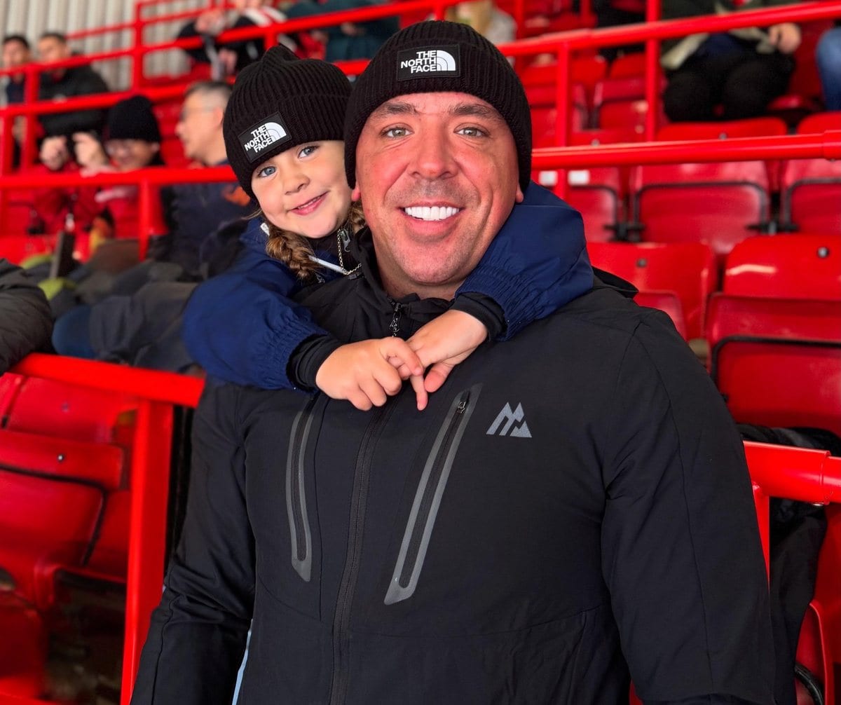 Charlie in a beanie at a football ground, a child on his shoulders, both smiling