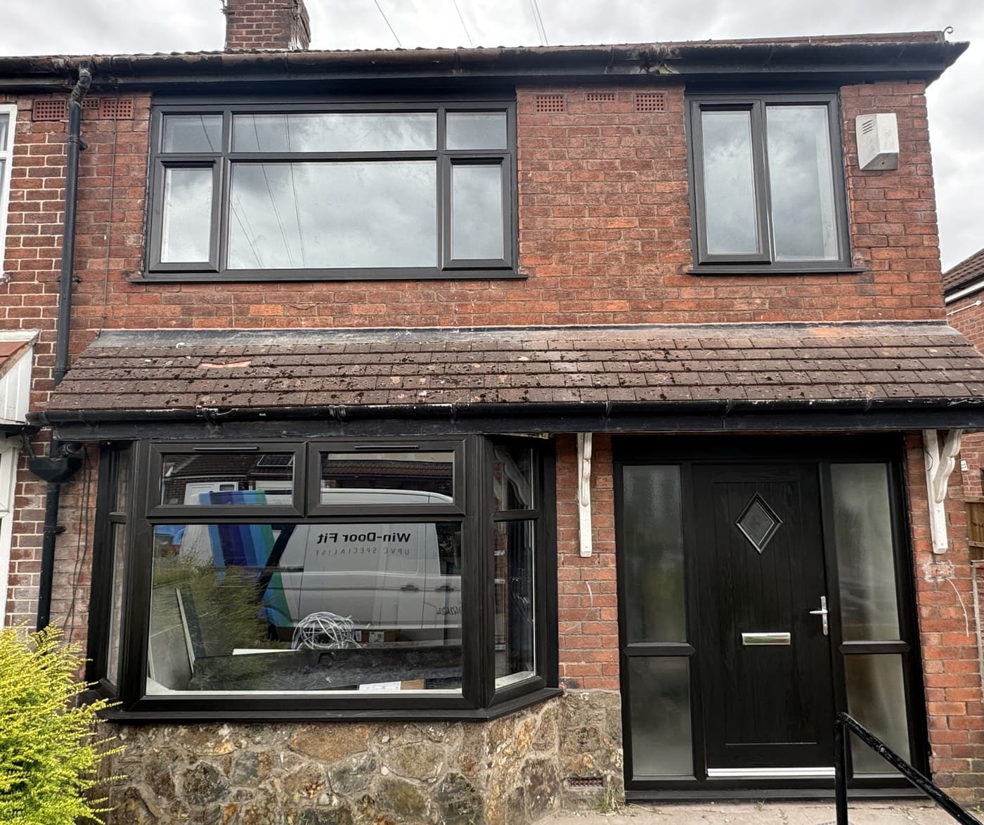 A red brick semi detached house with a new anthracite bay window and a black composite front door, the Win-Door Fit van reflected in the bay glass