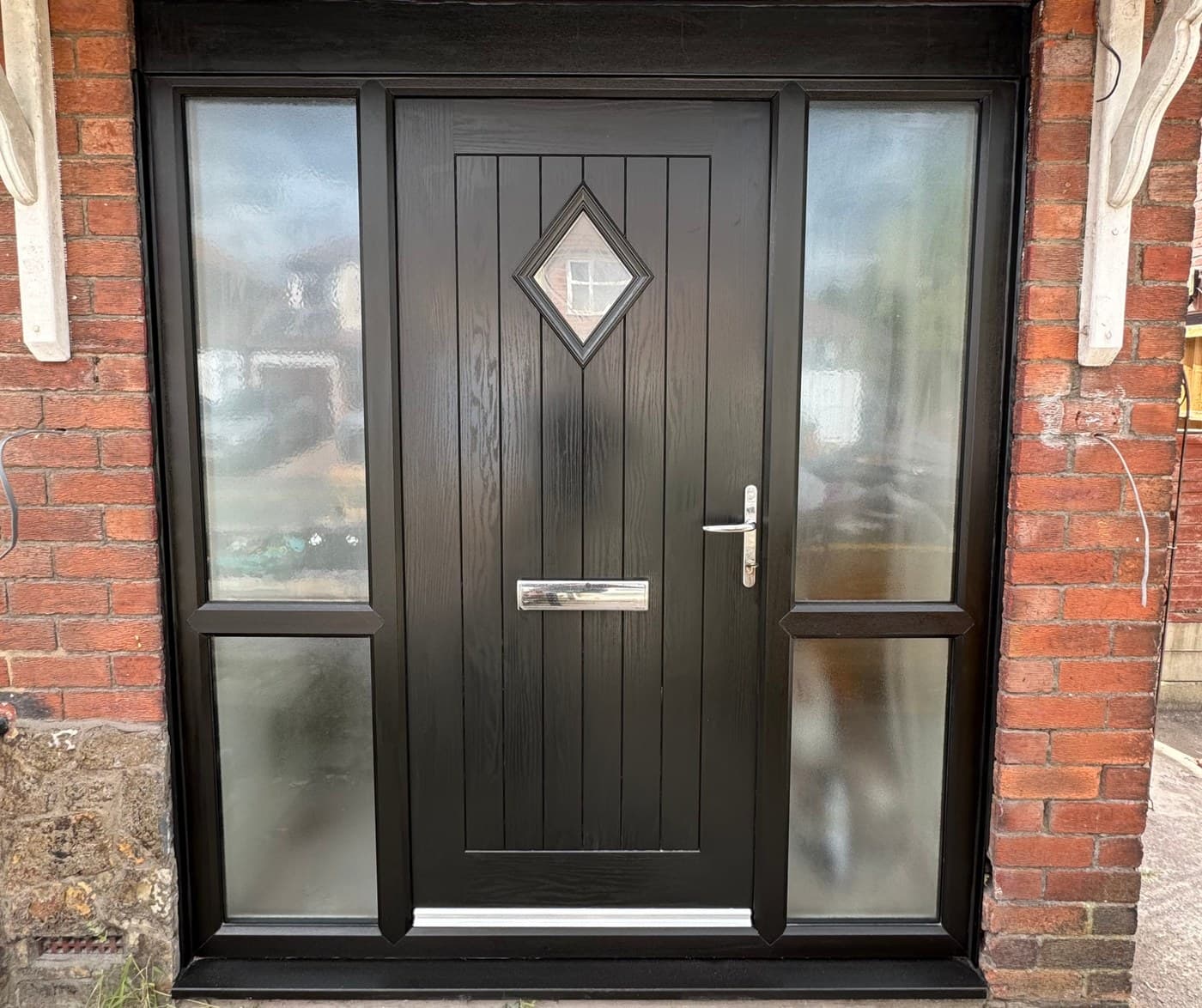 A dark composite front door with a diamond glazed panel and two glazed side panels, in a red brick surround