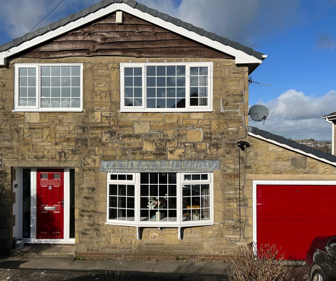A gritstone house with white uPVC bay windows and a red composite front door