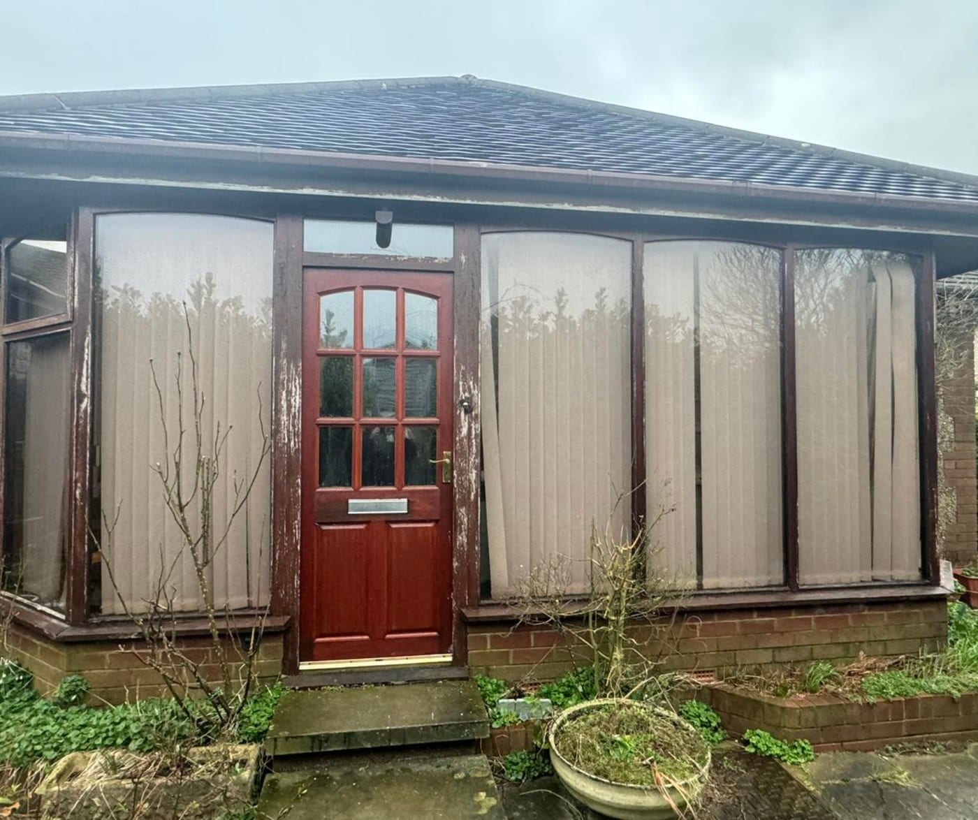 Before: a sunroom with rotted dark stained timber frames, net curtains and a red timber door with a nine pane top light