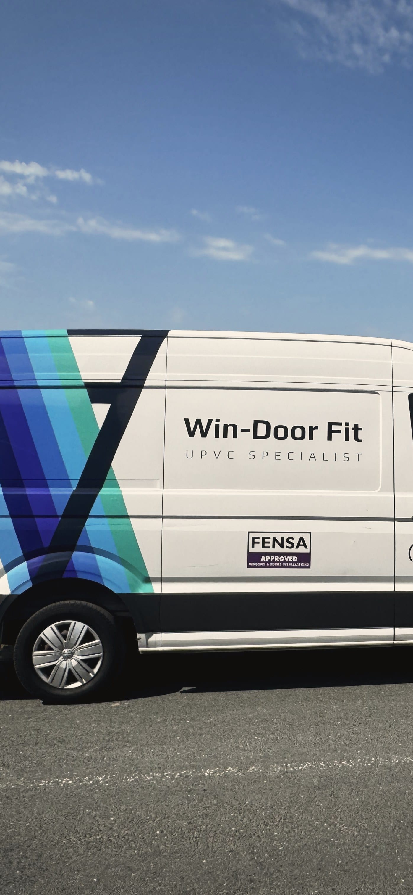 The Win-Door Fit van parked side on at the edge of a country road, signwritten with the Win-Door Fit name, the words uPVC Specialist and a FENSA Approved plaque, with a dry stone wall, open hills and clear sky behind it