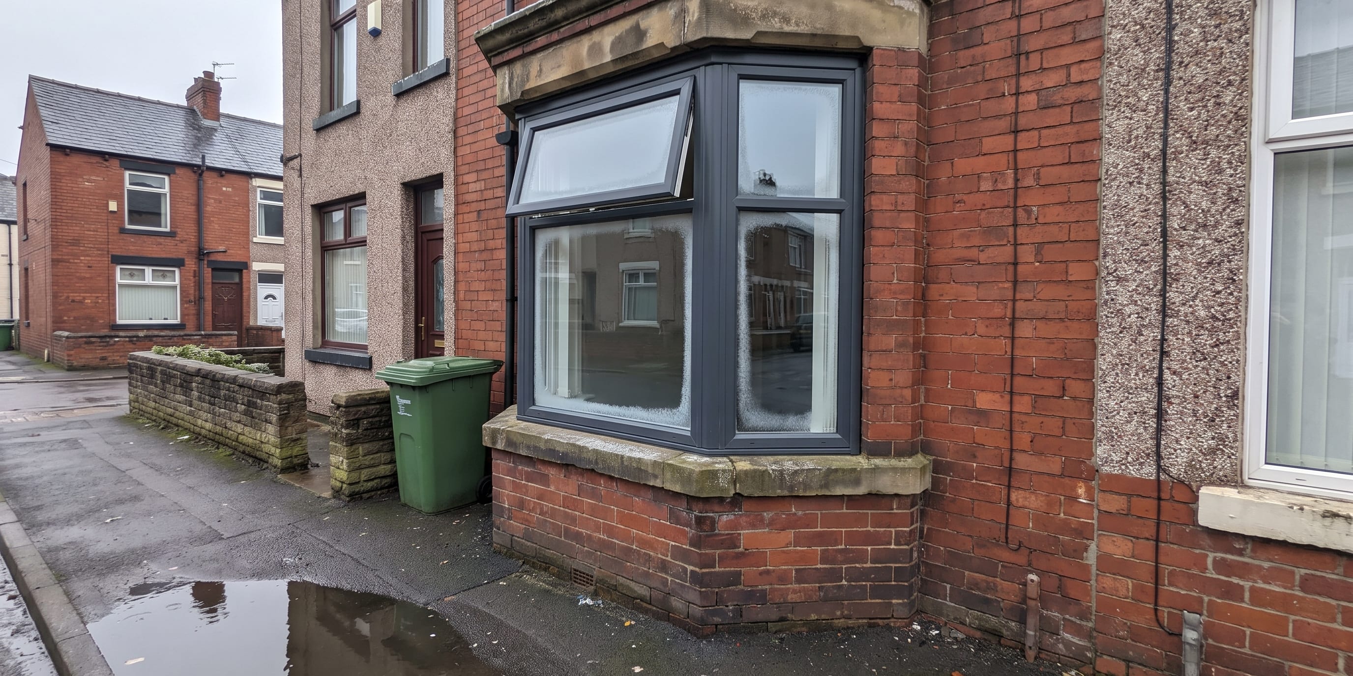 A red brick terraced house with a grey uPVC bay window fitted with a top-opening vent, next to a green wheelie bin on a wet street.