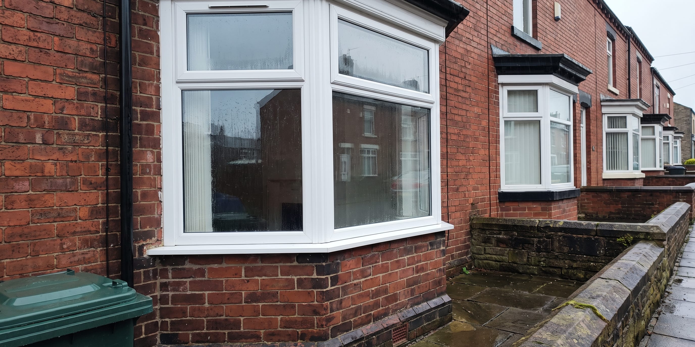 A white uPVC bay window on the front of a red brick terraced house, with a row of similar houses and a wheelie bin visible along a wet street.