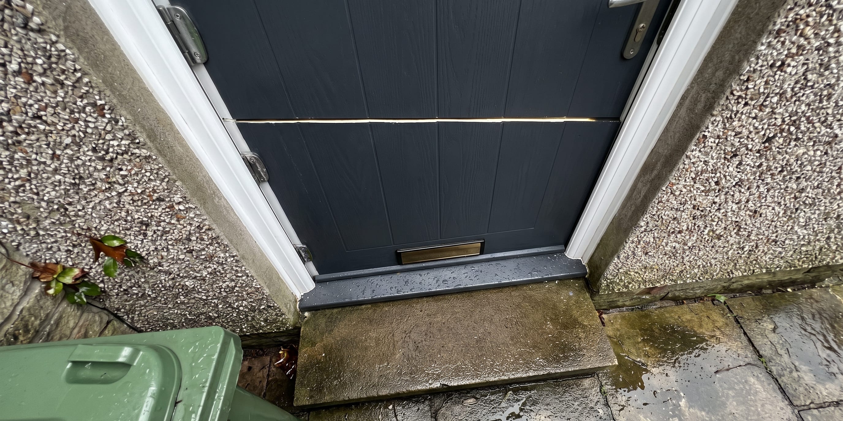 A dark grey composite front door viewed from above showing a thin horizontal line across its middle, with a stone step, wheelie bins and wet paving below.