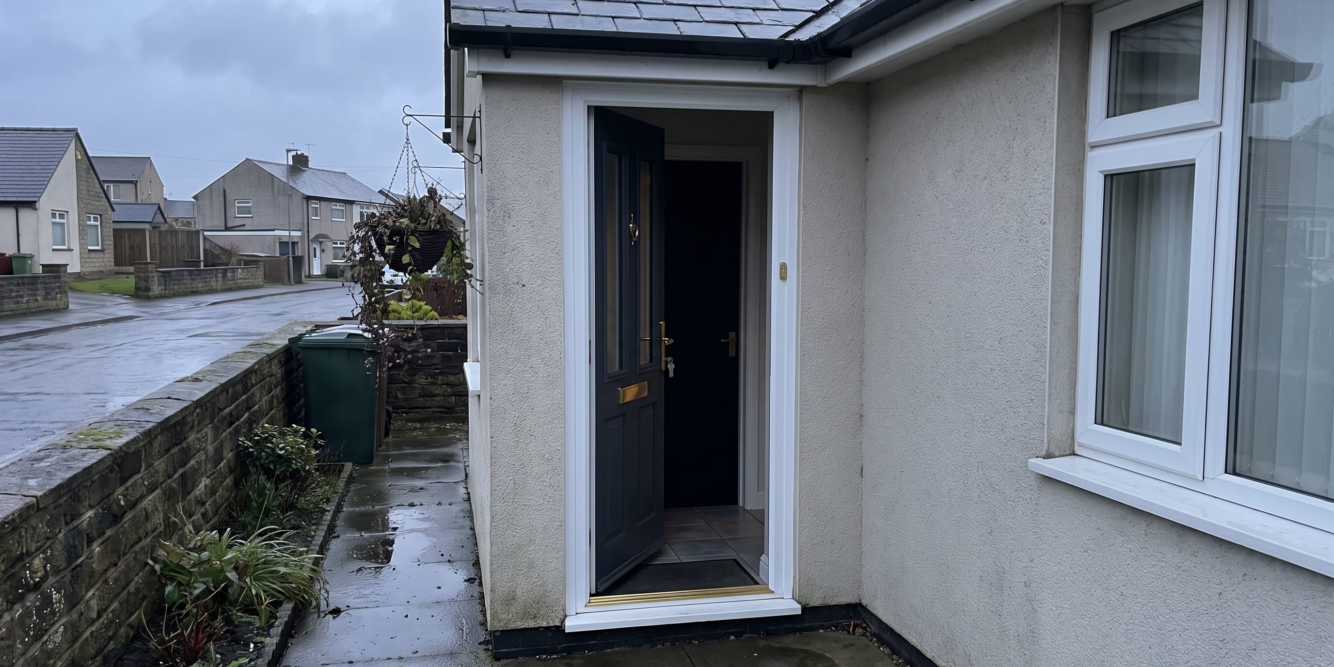 An open dark composite front door with a hanging basket beside it, set into a white uPVC frame on a rendered bungalow exterior.