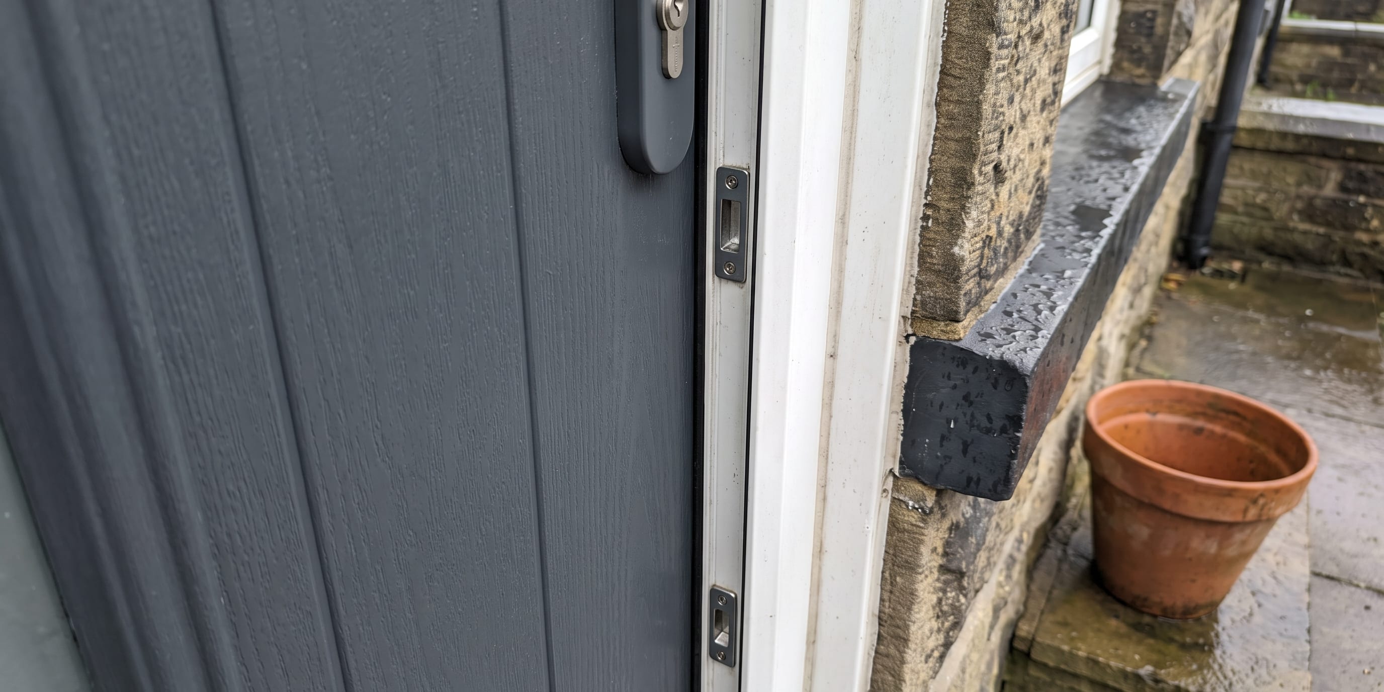Close-up of a grey uPVC door edge showing a cylinder lock and multi-point lock keeps set into a white frame beside a stone wall and terracotta plant pot.