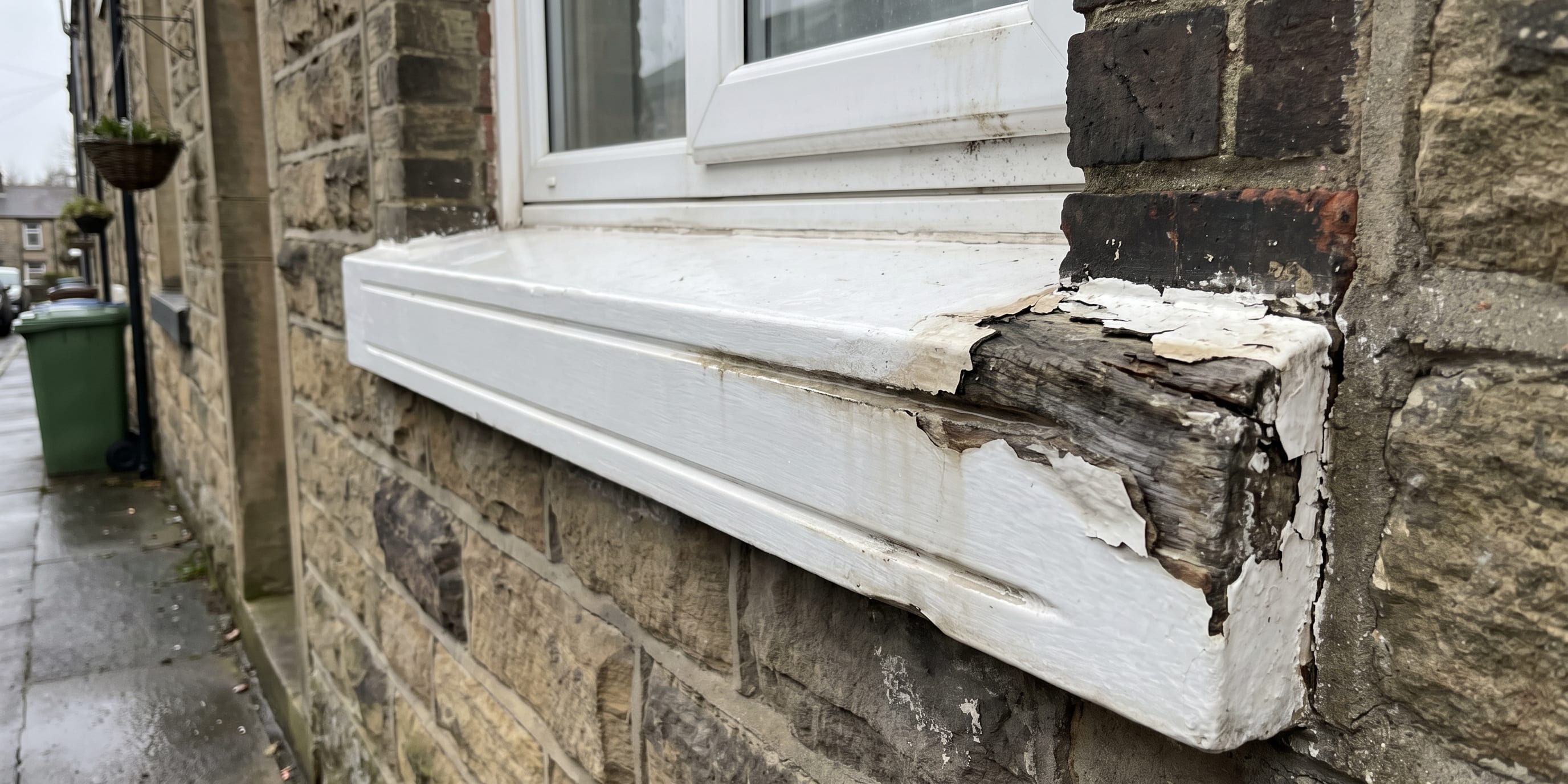 A white-painted timber window sill on a stone wall is badly rotted and peeling at the corner nearest the brickwork.